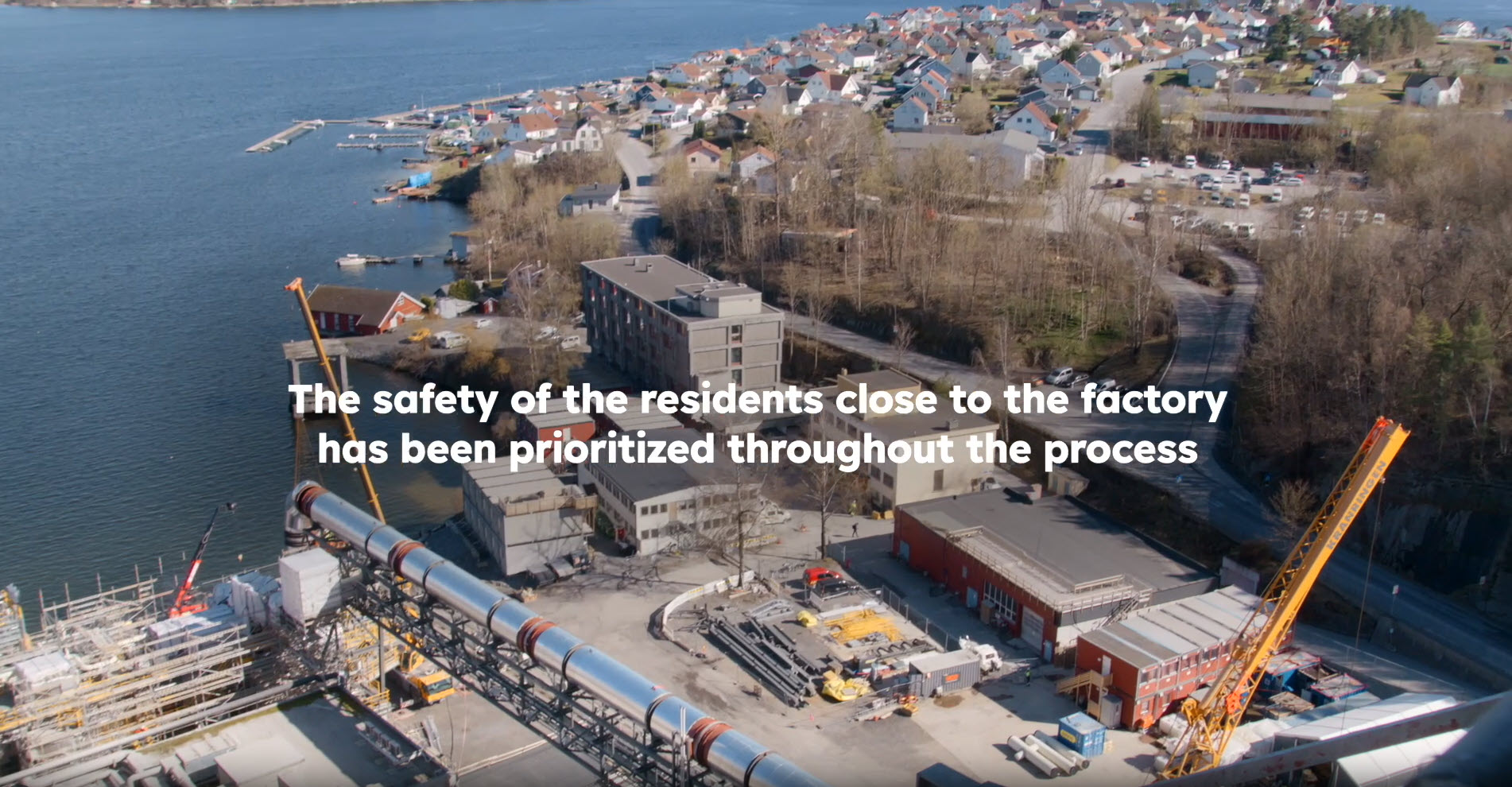 Aerial view of an industrial plant by the sea, behind it a village by the sea, above it the lettering "The safety of the residents close to the factory has been prioritized throughout the process."