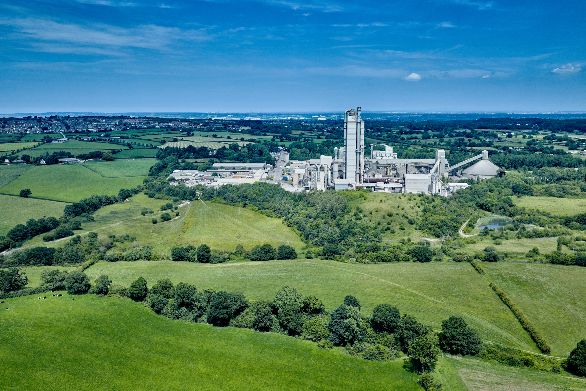 Aerial view of a large cement factory surrounded by green fields and trees, with industrial buildings and tall chimneys at the center. Rolling countryside extends into the background under a clear blue sky.