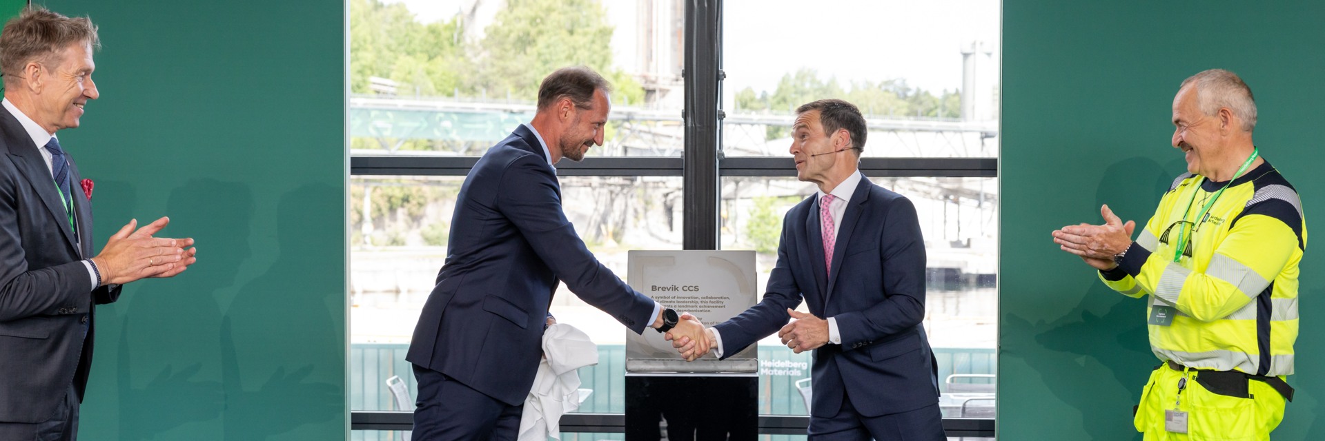 Two men in suits shake hands in the center, standing in front of a plaque and a window overlooking a greenery-lined bridge. On the left, a man in business attire claps, while on the right, a man in high-visibility workwear also claps. The event appears formal, celebrating a milestone or achievement.